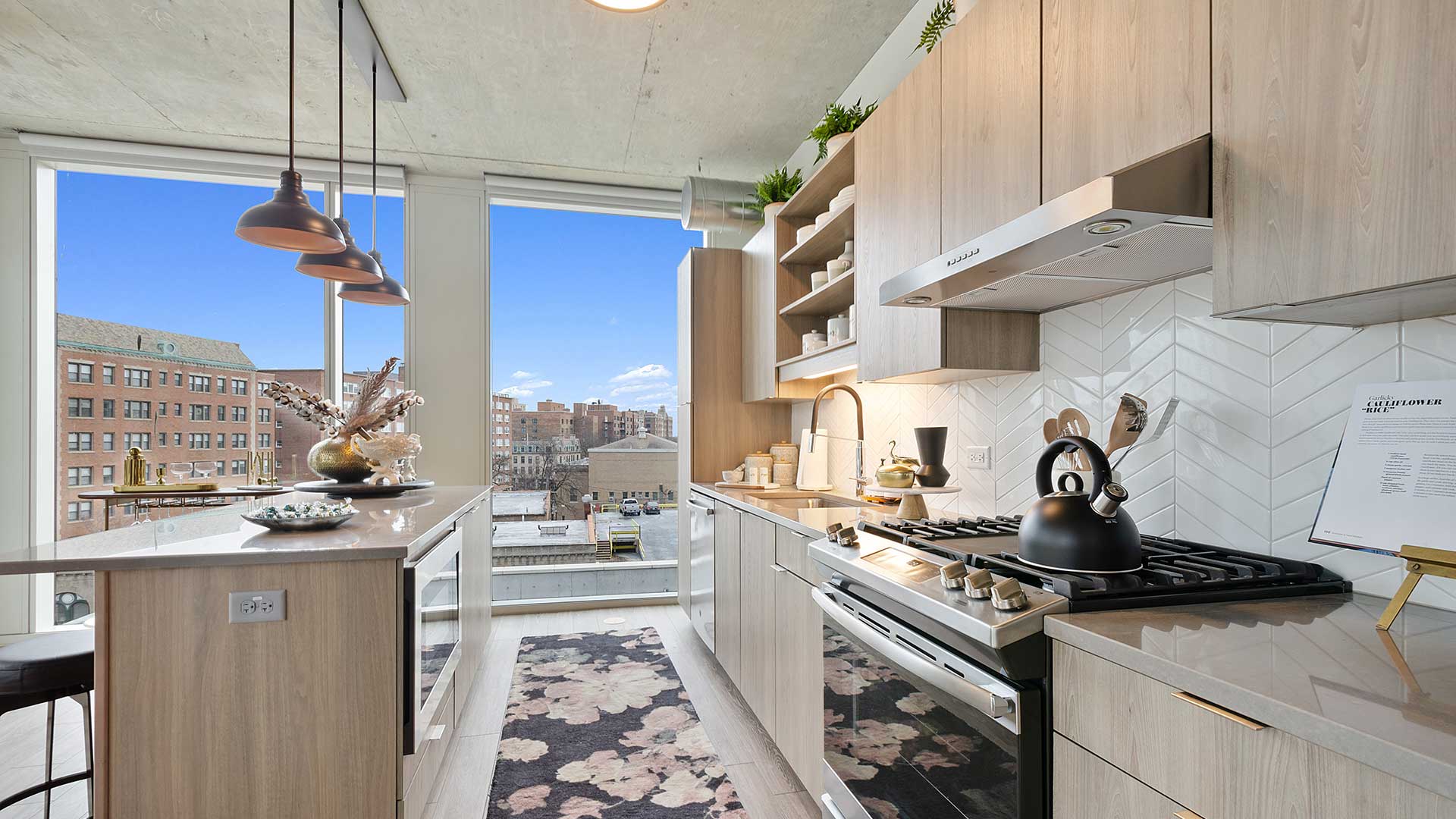 Contemporary apartment kitchen with quartz countertops, large island, stainless steel gas range, wood cabinetry, and floor-to-ceiling windows.
