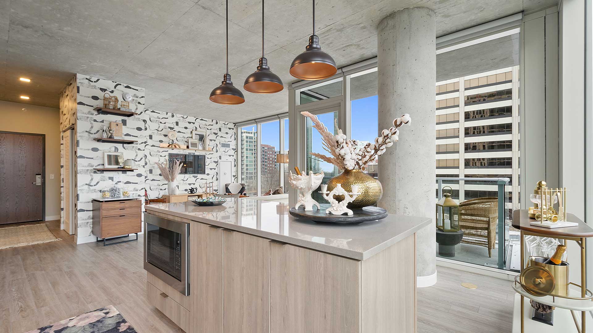 Open-concept apartment interior with large kitchen island, pendant lighting, floor-to-ceiling windows, and private balcony access.