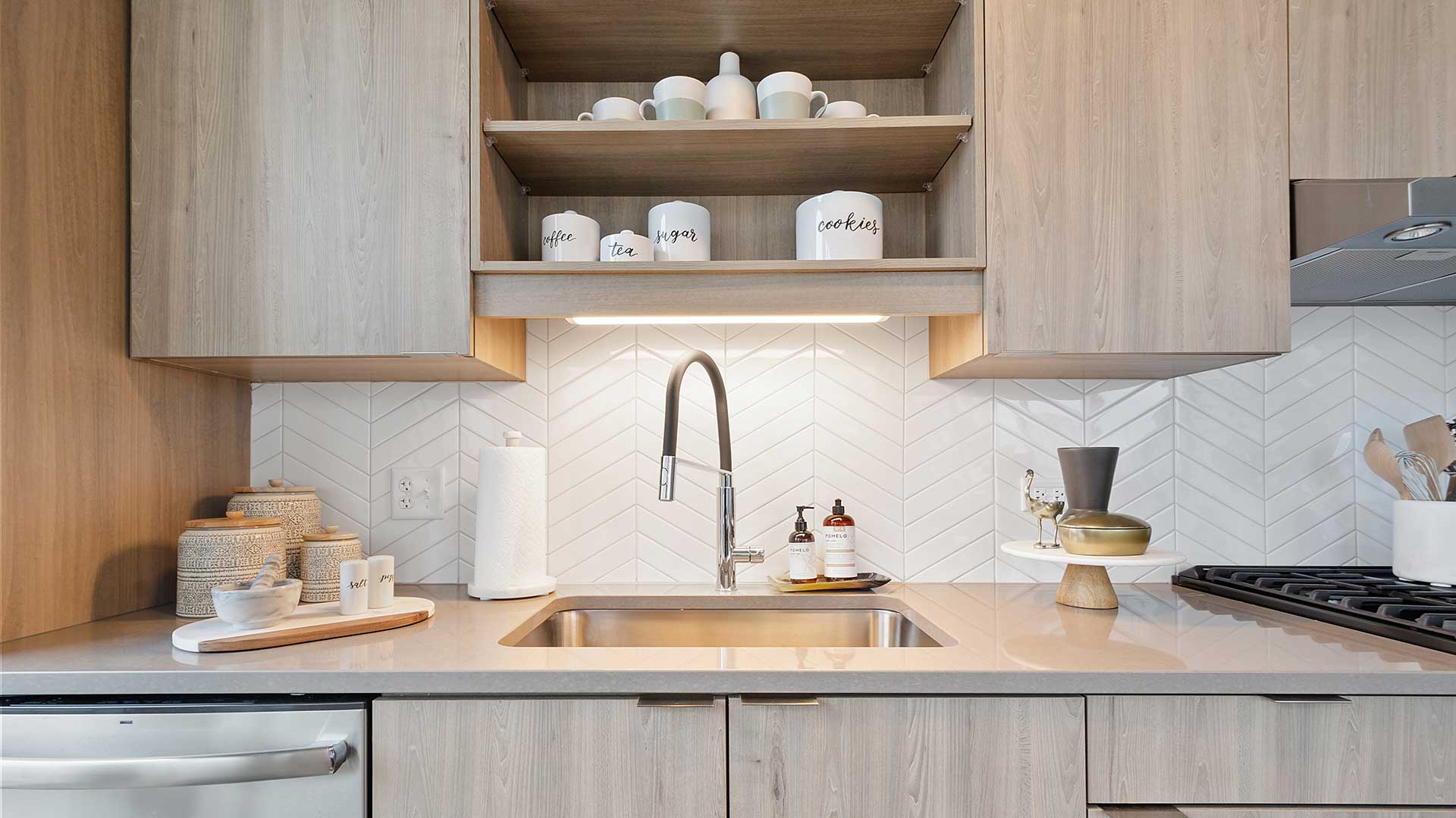 Close-up of apartment kitchen with wood cabinetry, open shelving, white chevron tile backsplash, quartz countertop, and modern faucet.