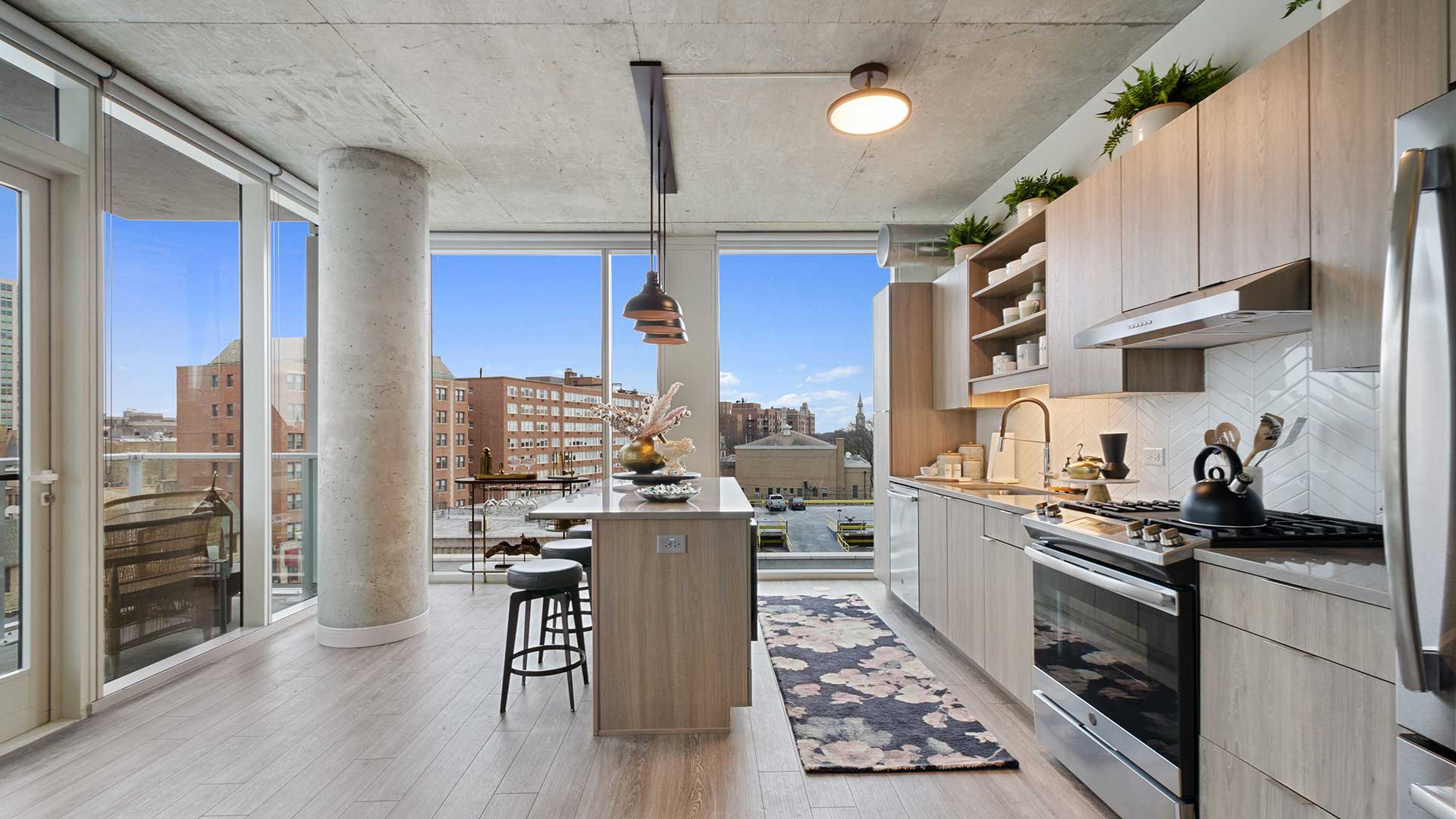 Modern apartment kitchen with center island, bar seating, wood cabinetry, stainless steel appliances, floor-to-ceiling windows, and balcony access.