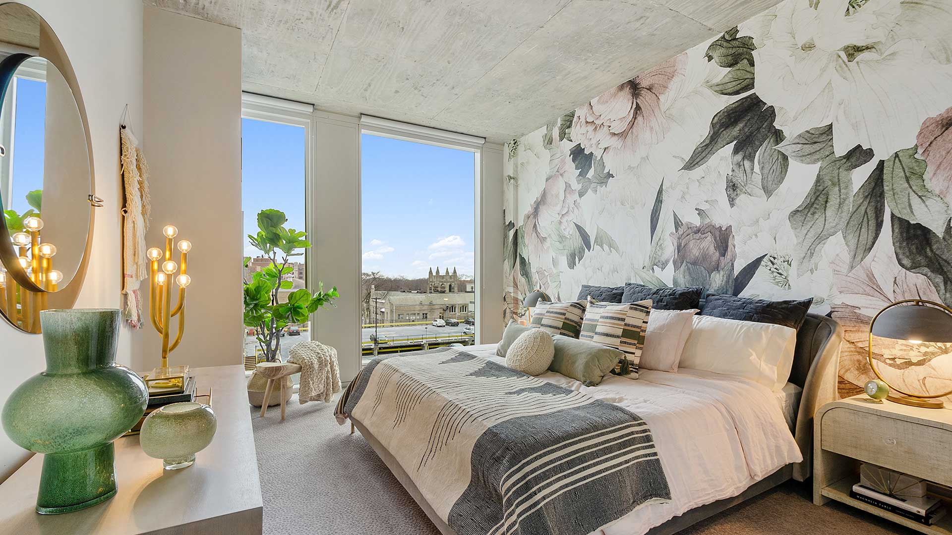 Modern apartment bedroom with floor-to-ceiling windows, a large floral accent wall, upholstered bed and contemporary furnishings.