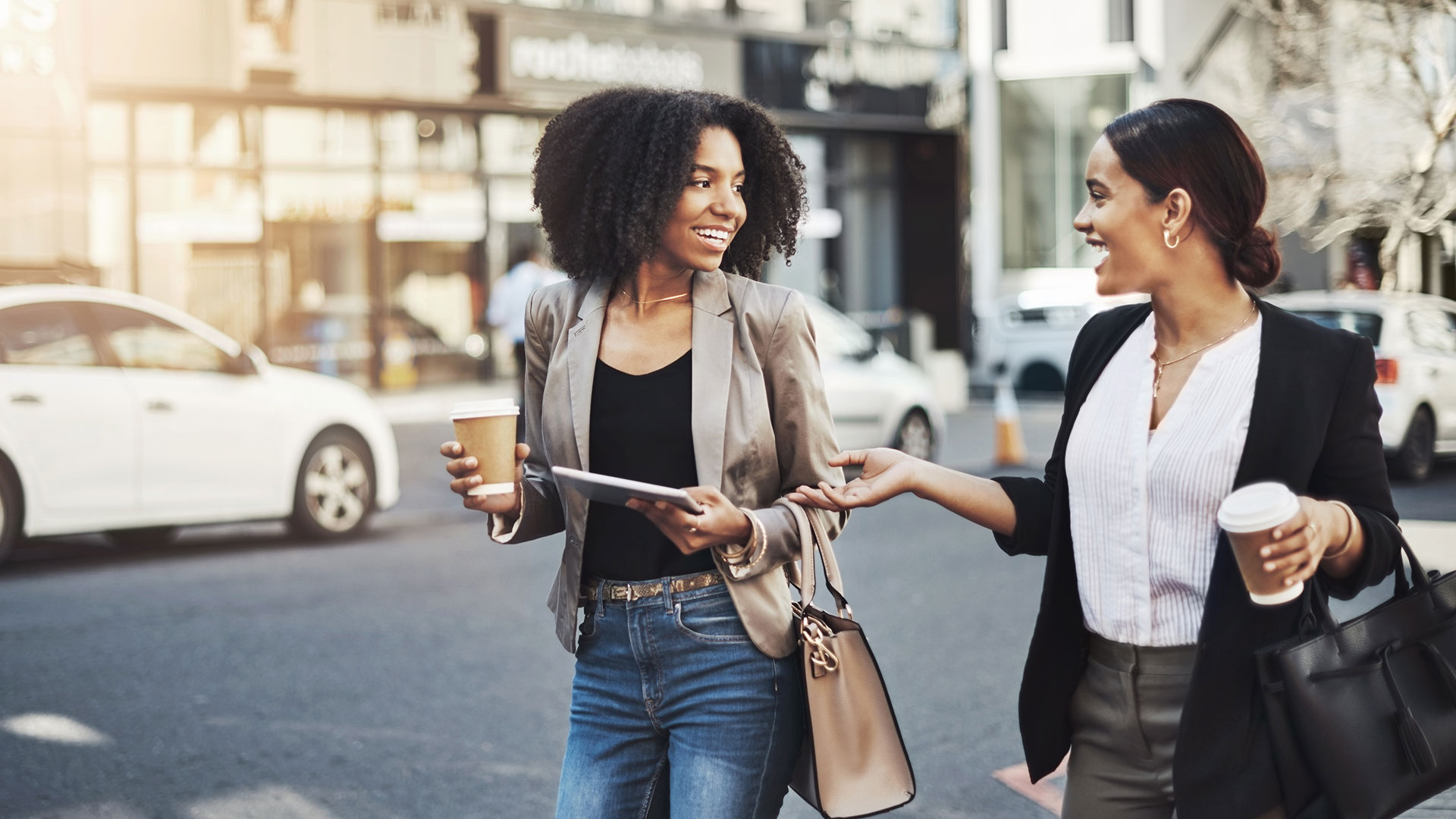 Two women walking through a city neighborhood with coffee and a tablet.